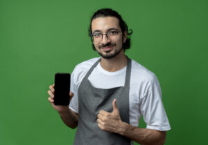 pleased young caucasian male barber wearing uniform and glasses holding mobile phone and showing thumb up isolated on green background